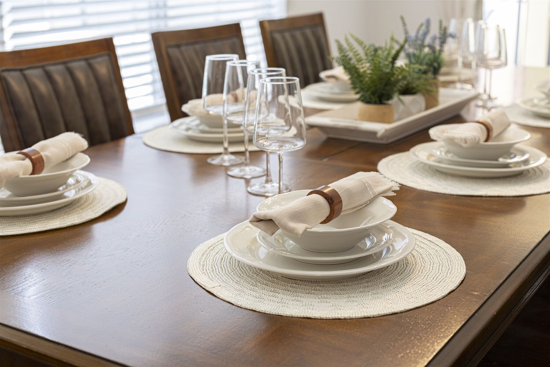 Una mesa de comedor dispuesta con platos, fuentes y vasos blancos, con servilletas dobladas sobre individuales.