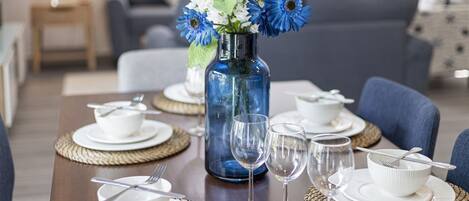 Una mesa de comedor dispuesta con cuencos, platos y copas de vino, junto a un jarrón con flores azules.