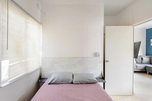 Ein Schlafzimmer mit einem Bett, einem Nachttisch, einem Fenster mit Jalousien und einer Tür zu einem anderen Raum.