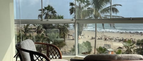 Un balcón con sillas de mimbre y una mesita, con vistas a una playa con palmeras y un cielo despejado.