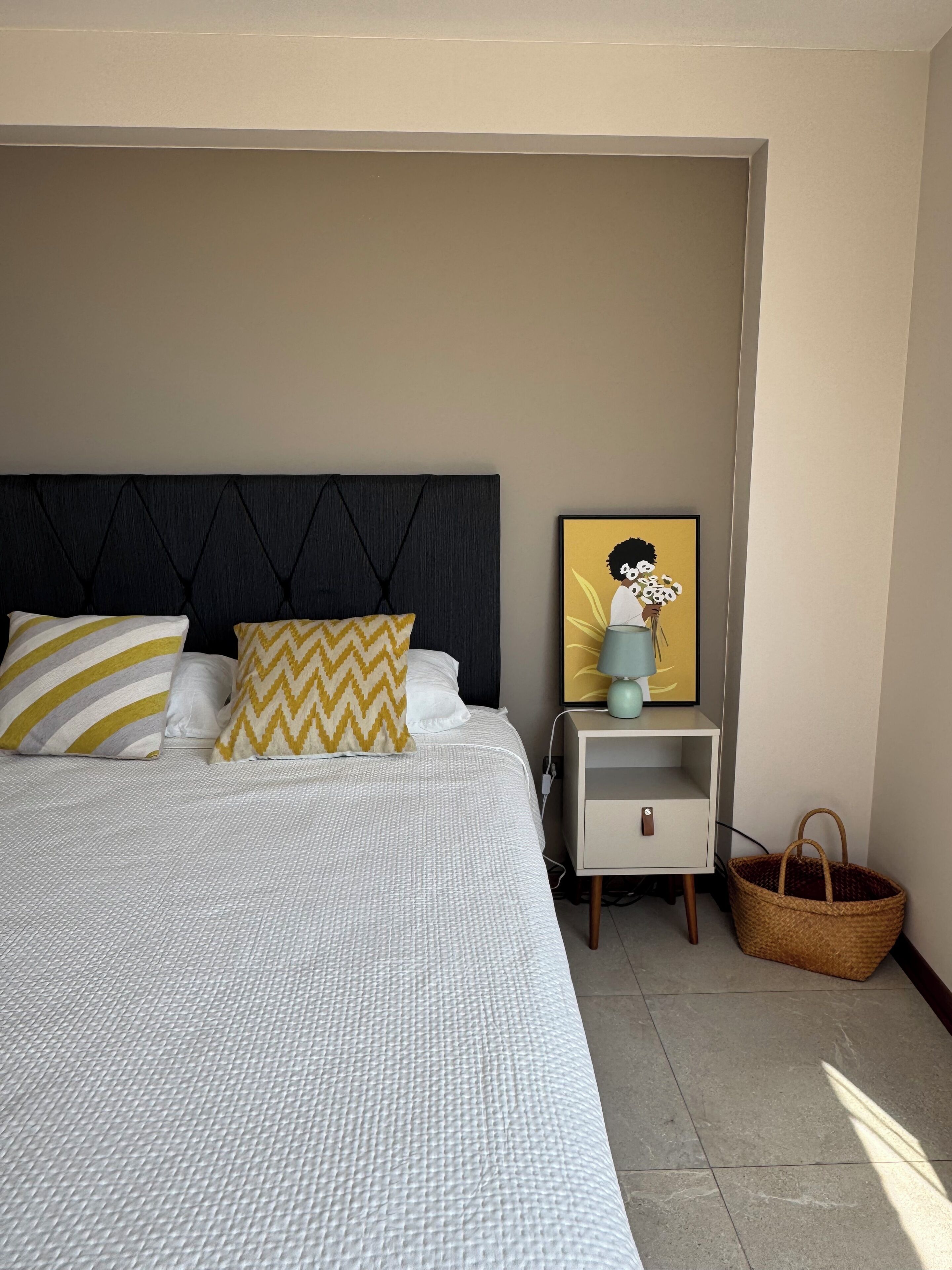 Un lit bien fait, avec une couvre-lit à motifs et deux oreillers, une table de chevet avec une lampe et un tableau encadré, et un panier en osier posé sur le sol.