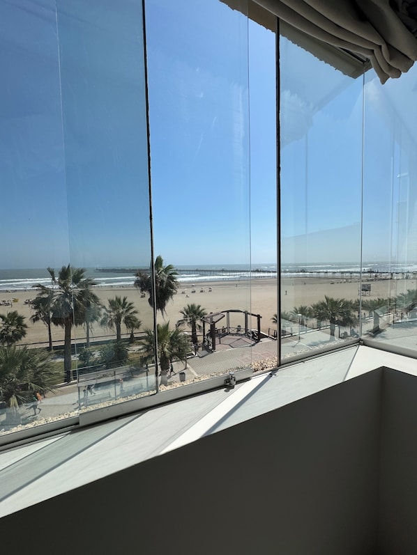 Vista desde una ventana hacia una playa con palmeras y un paseo marítimo.