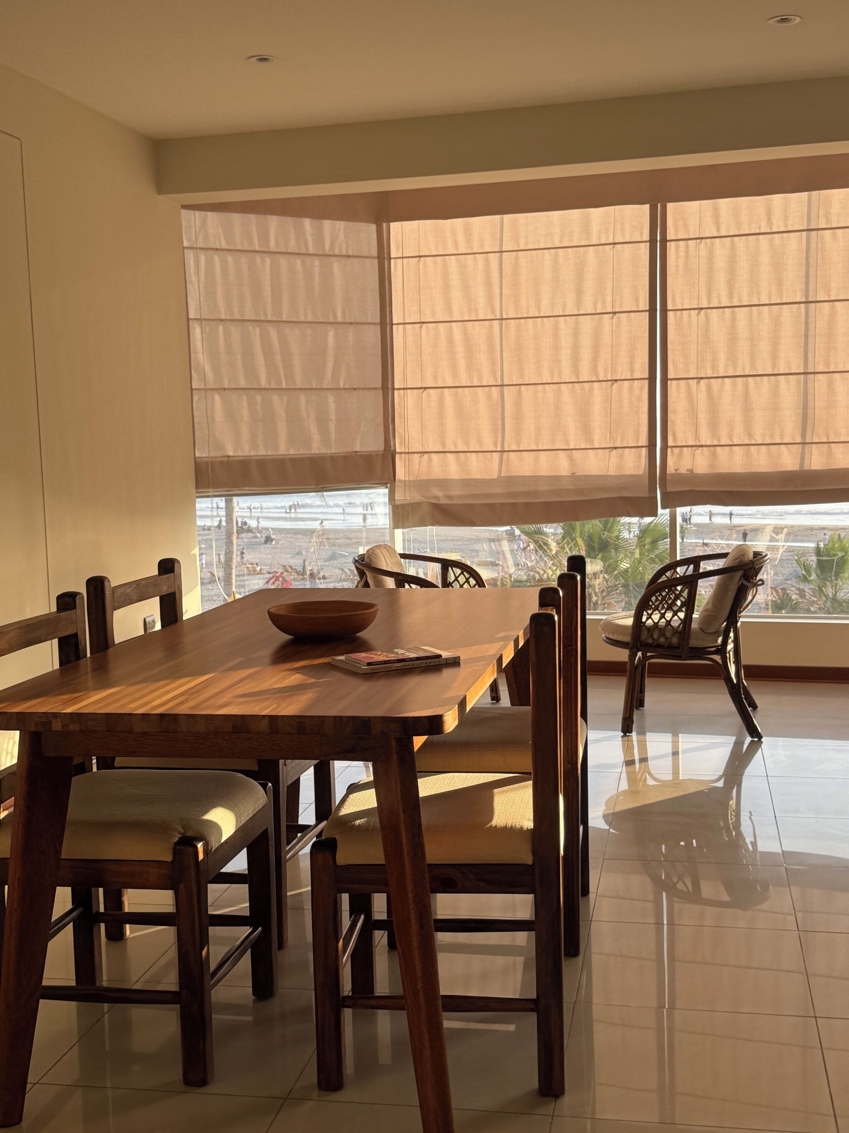 A dining area with a wooden table and chairs, large windows with curtains, and a view of the beach.