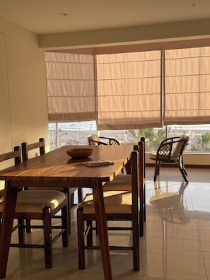 Un comedor con mesa y sillas de madera, ventanas amplias con cortinas y vista a la playa.