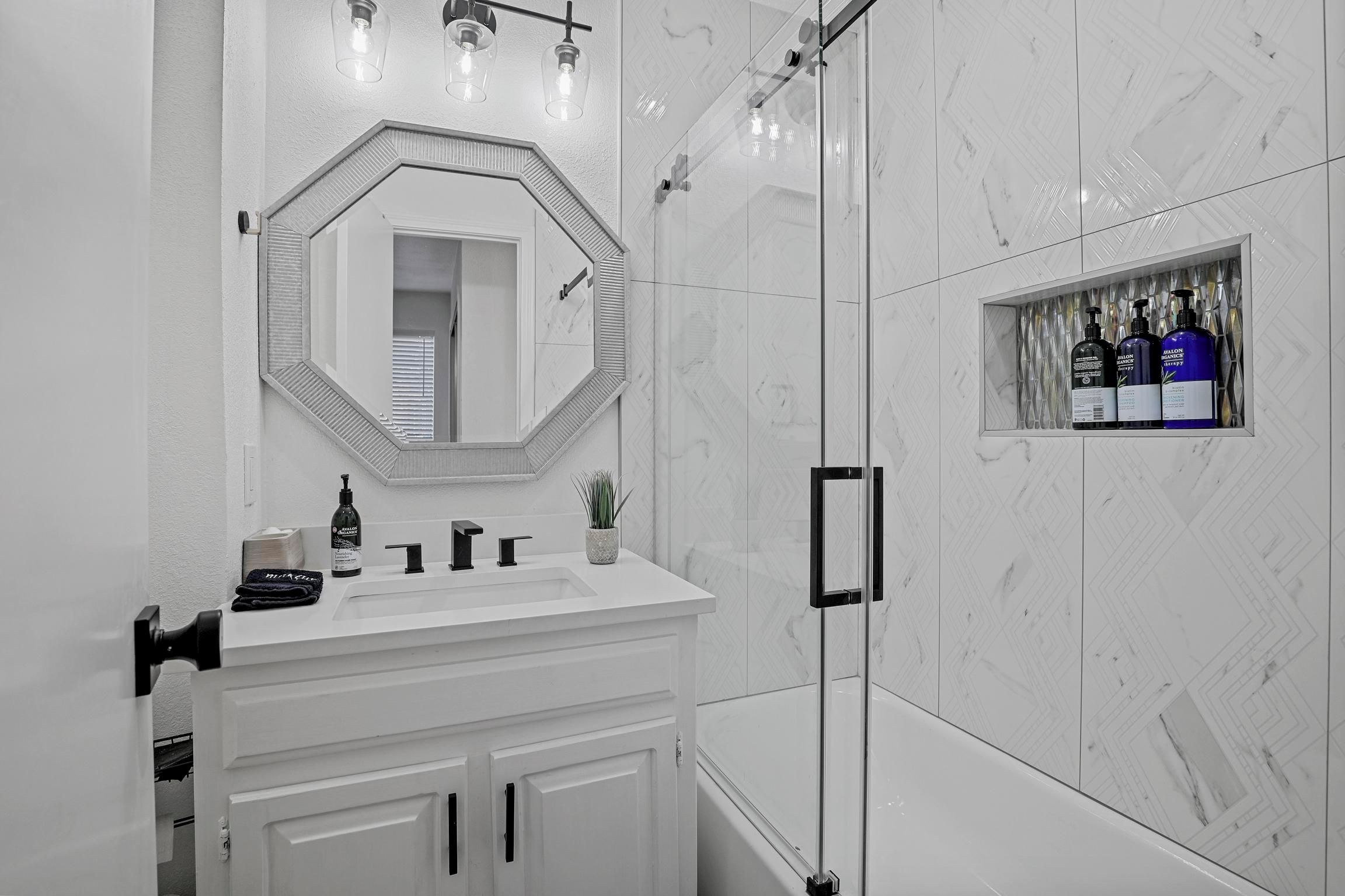 Une salle de bain avec un meuble-lavabo blanc, un grand miroir, un espace douche et une étagère contenant des articles de toilette.