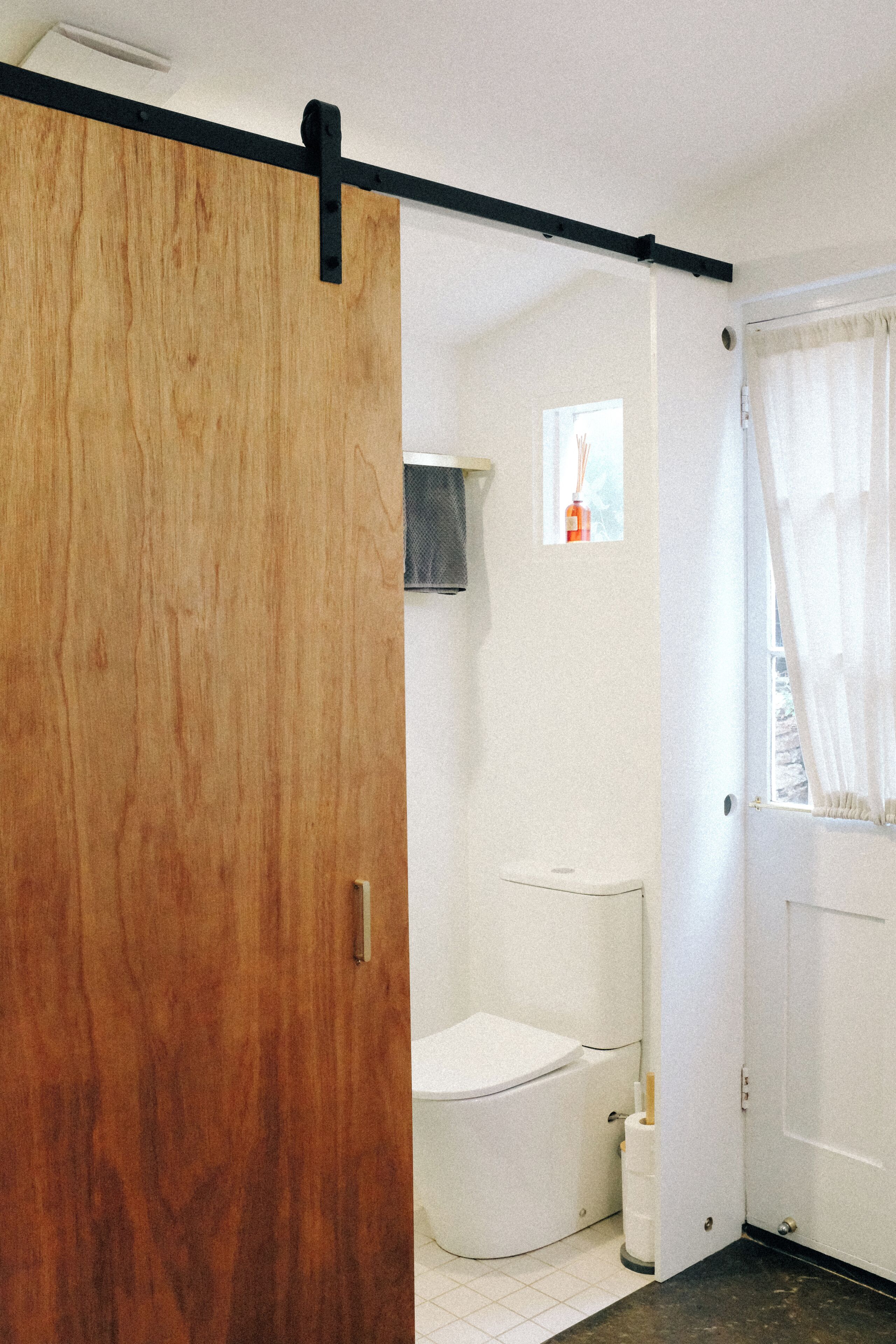 A bathroom with a wooden door, a toilet, a small window, and a shower area.