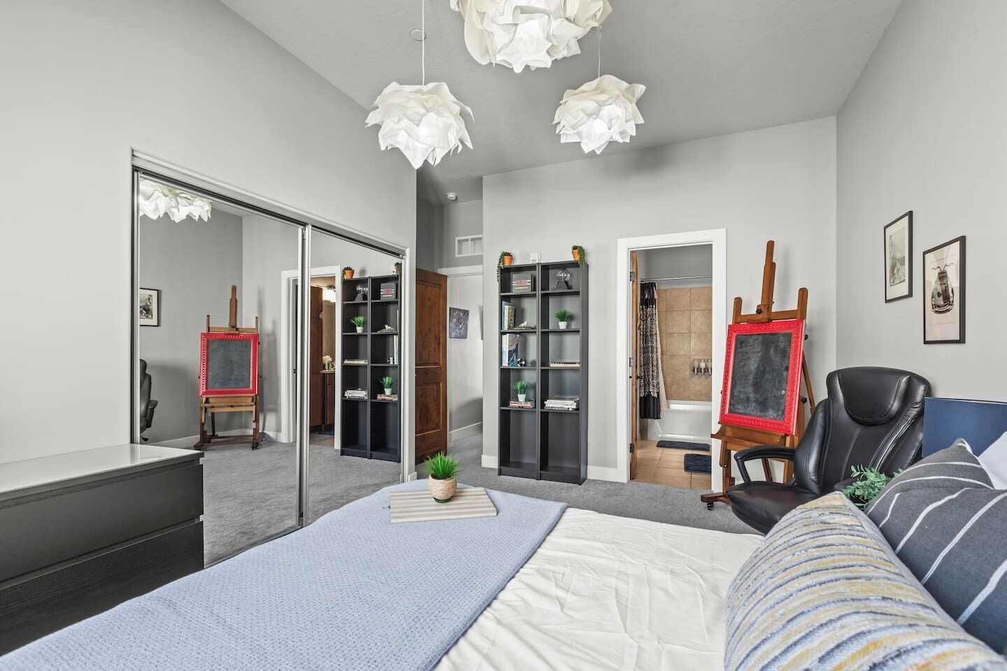 A bedroom with a bed, a desk, a chair, a bookshelf, and a bathroom visible through an open door.
