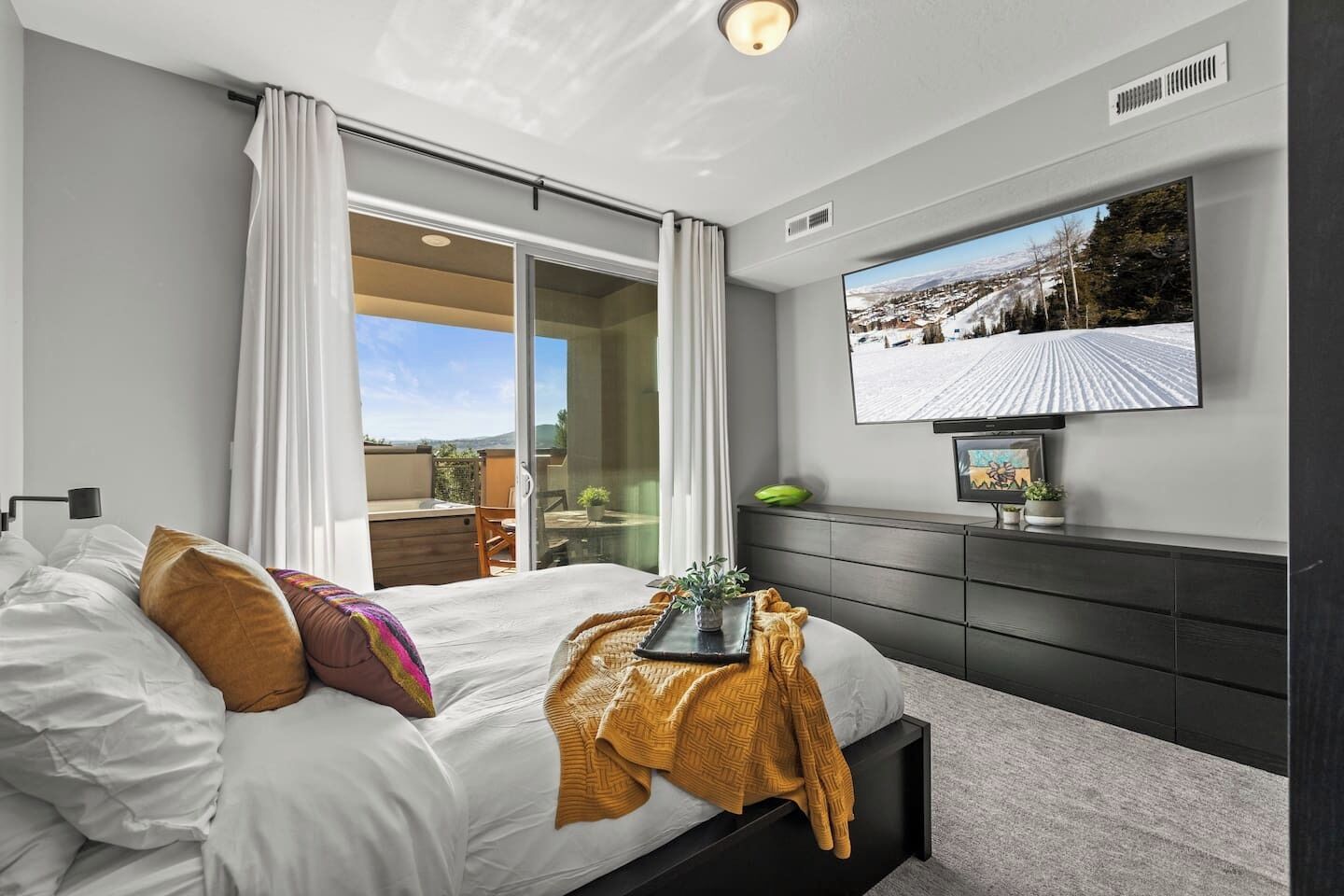 A modern bedroom with a bed, a flat-screen TV, a sliding glass door to a balcony, and a dark wooden dresser.