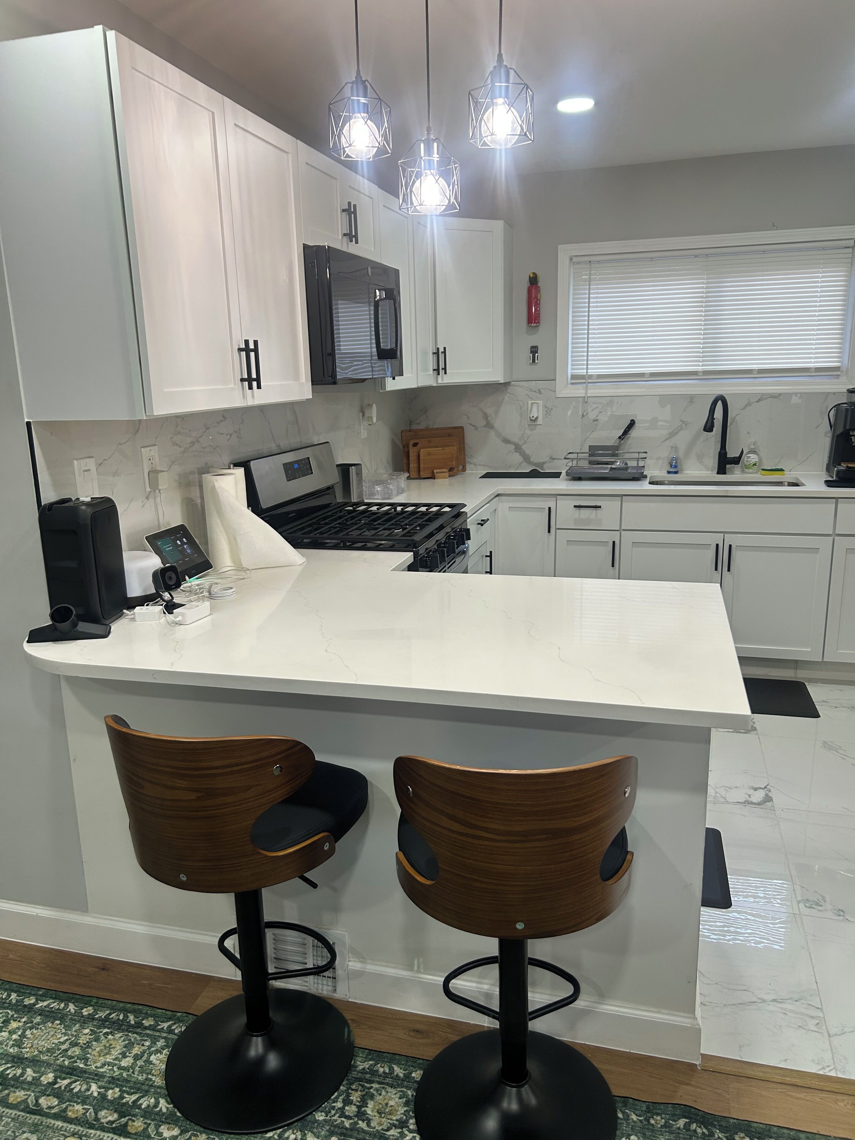 A modern kitchen with white cabinets, a marble countertop, a built-in oven, a sink, and a coffee maker. There are two wooden bar stools with black bases.