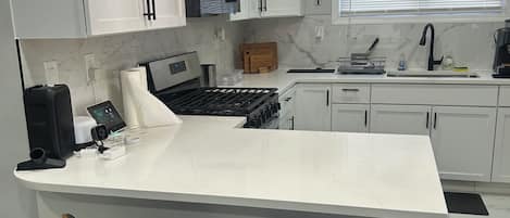 A modern kitchen with white cabinets, a marble countertop, a built-in oven, a sink, and a coffee maker. There are two wooden bar stools with black bases.