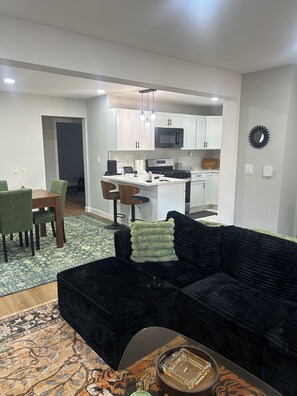 A modern living room with a black sofa, a dining area with green chairs, and a kitchen with white cabinets and a microwave. - Spacious 6-bedroom house with AC in vibrant Detroit (Detroit)