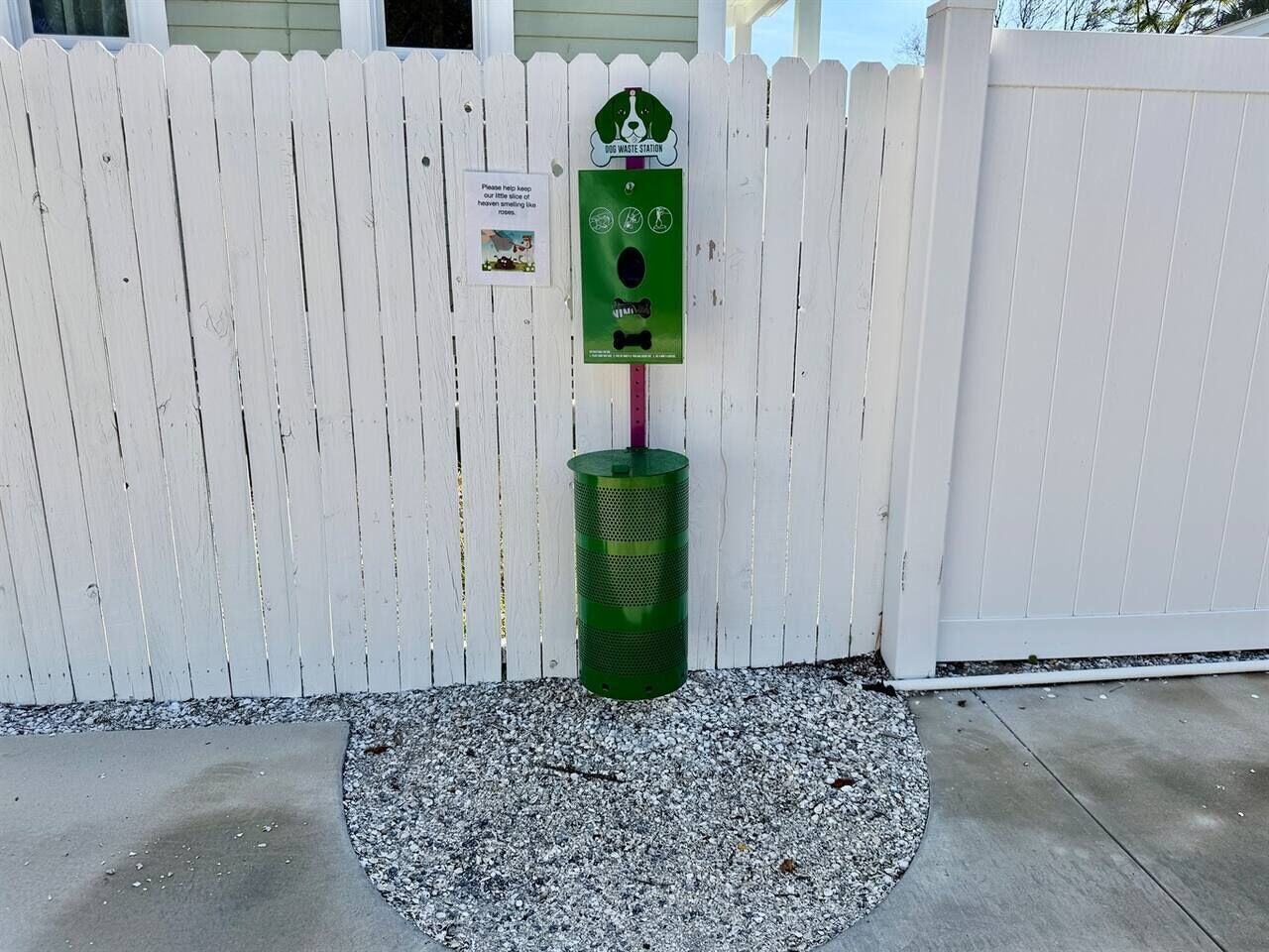 Un contenedor verde para reciclaje con un letrero, una cerca blanca de piquetes y un sendero de concreto.