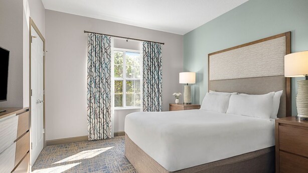 Habitación de hotel con una cama grande, una mesita de noche con lámpara, una ventana con cortinas y un televisor a la izquierda.
