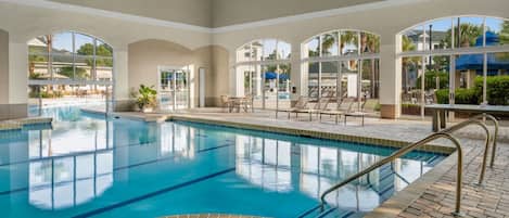 An indoor swimming pool with clear glass walls, lounge chairs, and a tiled floor.