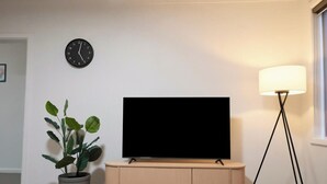 Ett rum med en tv på ett stativ, en klocka på väggen, en växt och en golvlampa.