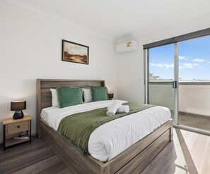 A bedroom with a bed, a nightstand, a lamp, a painting, and a sliding glass door to a balcony.