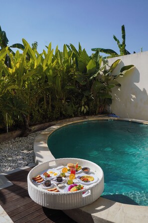 Un área junto a la piscina con una silla de mimbre, una mesa de comedor con alimentos para el desayuno y exuberante follaje tropical.
