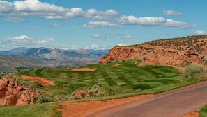 A golf course with a road curving around it, surrounded by red rock formations and distant mountains.