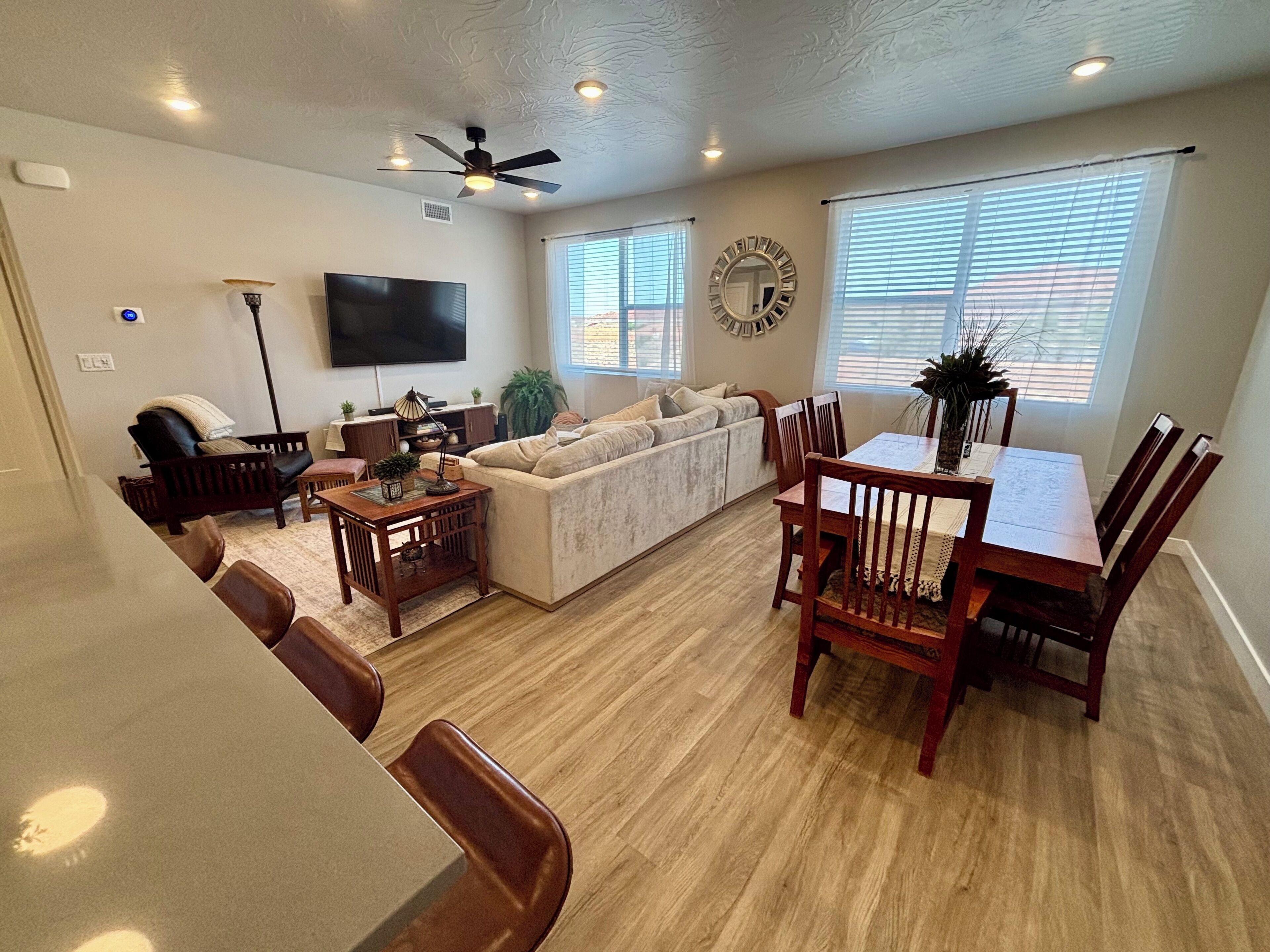 A living room with a sofa, TV, ceiling fan, dining table with chairs, and large windows.
