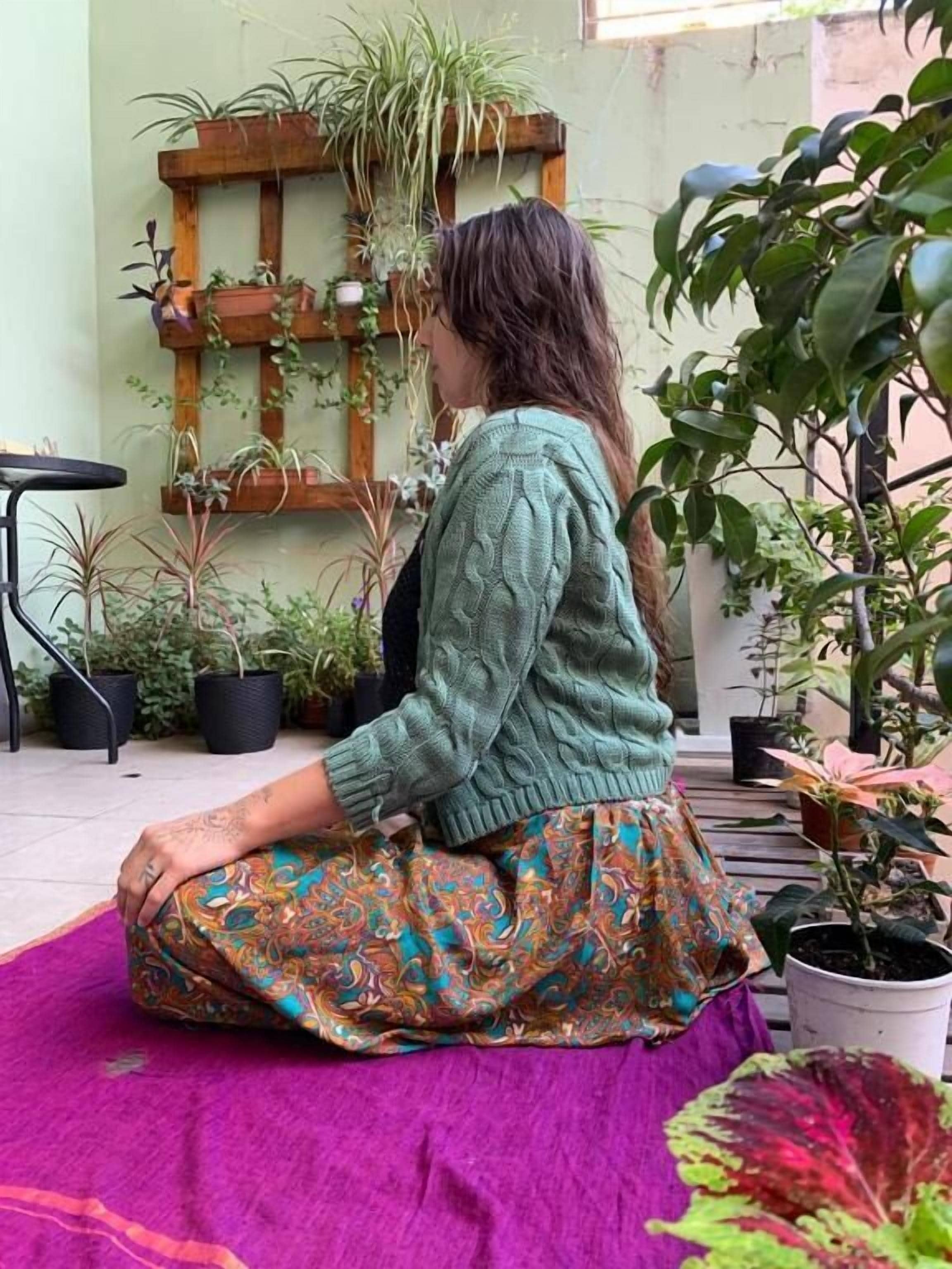 Una persona sentada sobre una esterilla morada en un espacio al aire libre con plantas en macetas y una estructura de madera.