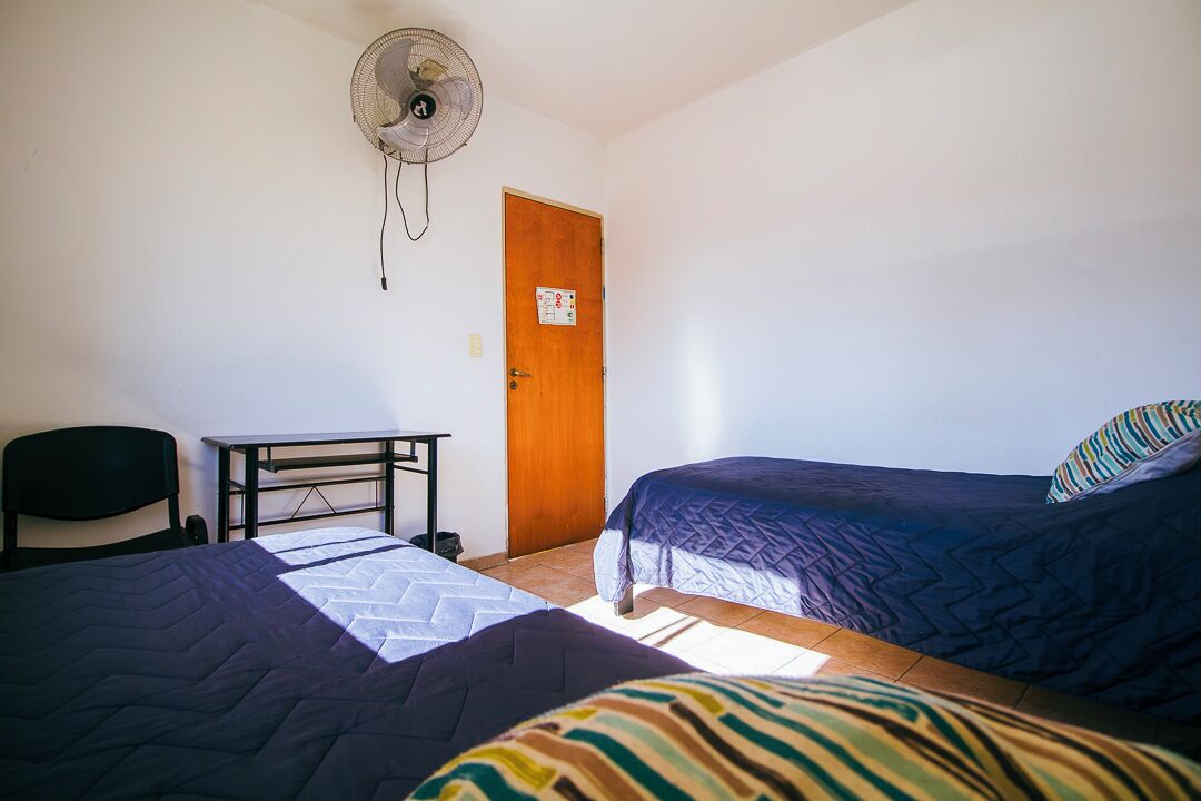 A room with two beds, a desk, a chair, and a standing fan.