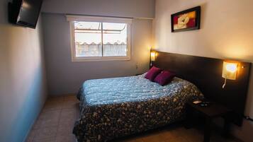 Un dormitorio con cama, mesita de noche, televisor en la pared, ventana y lámpara de pared.