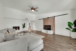 A spacious living room with a white brick fireplace, a large grey sofa, a coffee table, and a TV on a wooden stand. - Spacious Entire Home •4BR  •7 Beds •Sleeps 12+ • Near Dallas, Plano, Richardson (Garland)