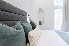 A bedroom with a bed, pillows, a headboard, a bedside table, a mirror, and a window.