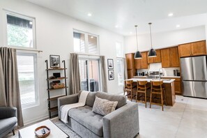 Una sala de estar moderna con un sofá gris, una mesita de centro y una cocina con armarios de madera y electrodomésticos de acero inoxidable.