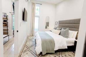 A bedroom with a bed, a bedside table, a mirror, a window with curtains, and a carpeted floor.