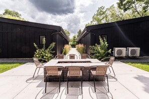 An outdoor dining area with a long table and chairs, flanked by two black buildings, under a cloudy sky.