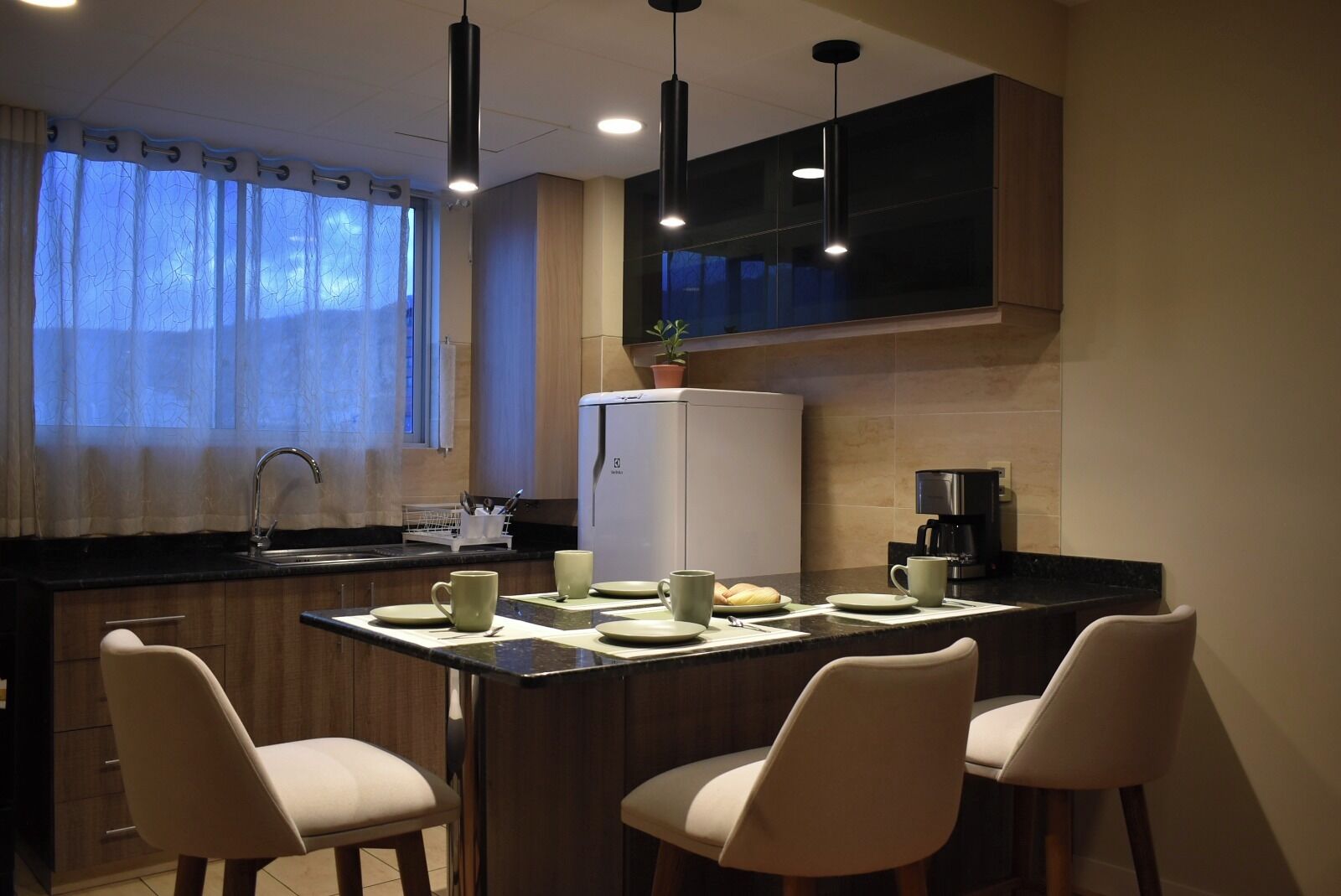 A modern kitchen with a dining area, including a refrigerator, coffee maker, and bar stools.