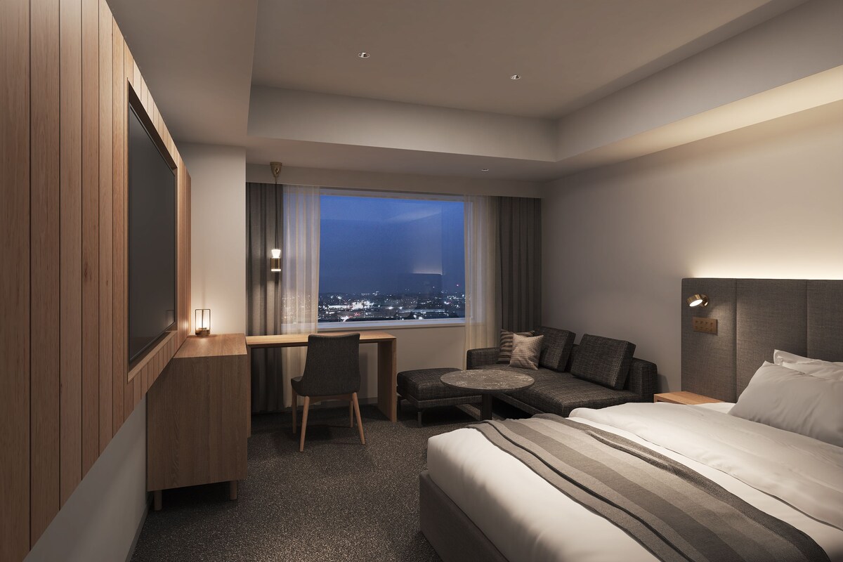 A modern hotel room with a bed, a desk, a chair, a sofa, and a view of the city at night.