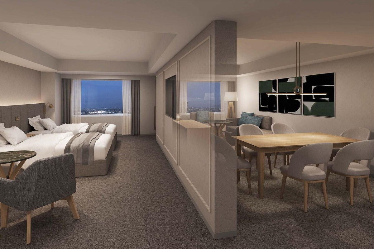 A hotel room with a bed, a desk, a dining area, and a view of the city.
