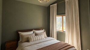 A bedroom with a bed, a nightstand, a hanging light fixture, a window with curtains, and a wooden ceiling beam. - Château Kozy Kove (Kawartha Lakes)