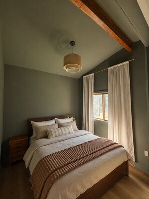 Ein Schlafzimmer mit einem Bett, einem Nachttisch, einer Hängeleuchte, einem Fenster mit Vorhängen und einem Holzbalken an der Decke.