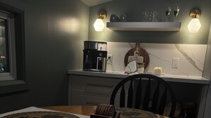 A dining area with a round table, placemats, a coffee maker, and a wall-mounted shelf with glasses. - Château Kozy Kove (Kawartha Lakes)