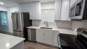 Una cocina moderna con armarios blancos, electrodomésticos de acero inoxidable, un lavamanos doble y un ventanal con persianas.