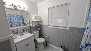Un baño con inodoro, lavamanos, espejo, toallero y una ventana con persianas.