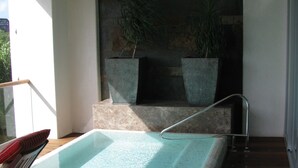 A pool area with a wooden deck, a seating area with a chair and a red cushion, and two large planters with plants. - Vidanta Golf and Spa Resort 1 bedroom Grand Luxxe suite (Playa del Carmen)