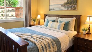A bedroom with a bed, two bedside tables with lamps, a window with curtains, a painting on the wall, and a wooden bench.