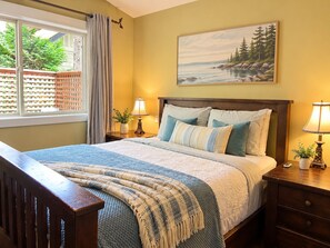 A bedroom with a bed, two bedside tables with lamps, a window with curtains, a painting on the wall, and a wooden bench.