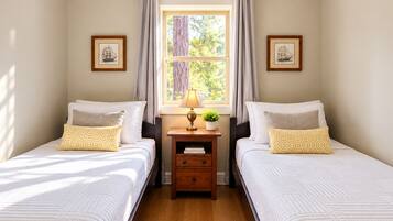 Una habitación con dos camas, una ventana con cortinas, una mesita de noche con lámpara y planta, y cuadros en las paredes.