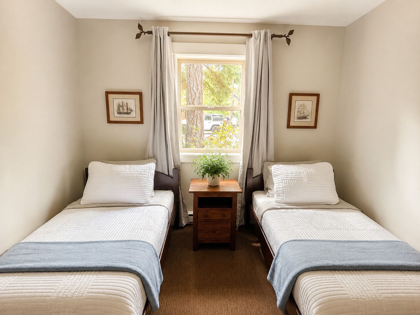 Una habitación con dos camas, una mesita con una planta y un ventanal con cortinas.