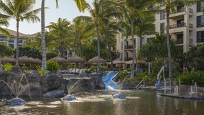 Zwembadgedeelte van een resort met palmbomen, ligbedden en een glijbaan.