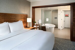 Hotelkamer met een groot bed, een houten hoofdbord, een nachtkastje met een lamp, een kleine plant en uitzicht op de badkamer met douche en bad.