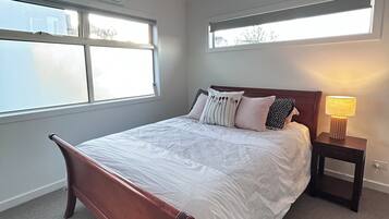 A bedroom with a wooden bed, white bedding, a nightstand with a lamp, and large windows.