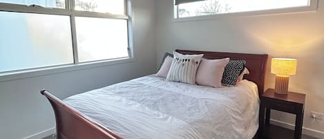 Un dormitorio con cama de madera, ropa de cama blanca, una mesita de noche con lámpara y ventanas amplias.