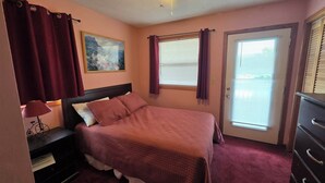 A bedroom with a bed, a nightstand, a lamp, a window with curtains, a door, and a ceiling fan. - Ocean & sound views, hot tub, in Nags Head. (Nags Head)