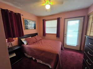 A bedroom with a bed, a nightstand, a lamp, a window with curtains, a door, and a ceiling fan. - Ocean & sound views, hot tub, in Nags Head. (Nags Head)